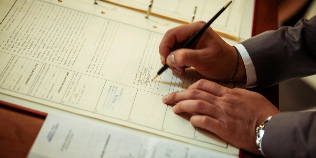 Mãos de um executivo de terno assinando um registro em um livro de documentos.