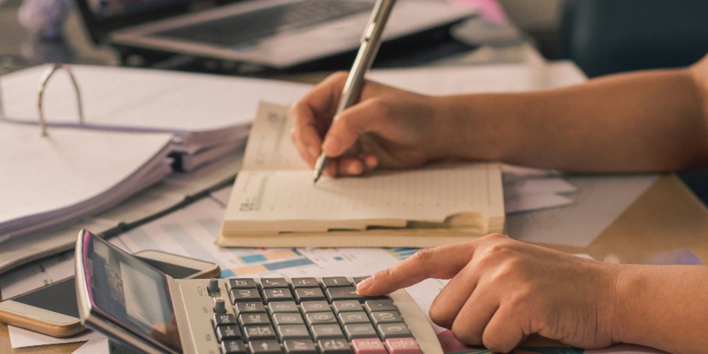Mãos fazendo contas em uma calculadora e anotando em um caderno sobre uma mesa com papéis.